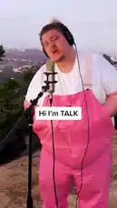 A passionate performance of 'How to Save a Life' by TALK, captured in a scenic outdoor setting. The artist, wearing pink overalls and headphones, sings into a professional microphone with heartfelt emotion. On-screen text introduces the performer and the song, creating an intimate and moving experience. by @iamtalk with iamtalk by Contains: How To Save A Life - TALK