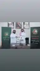 This official ceremony captures a key moment for Saudi Arabia's Al-Rawdha Club as two representatives formally sign an agreement during a press event in Riyadh. Dressed in traditional thobes and ghutras, they hold signed documents before branded banners displaying 'Al-Rawdha Club' and 'Riyadh'. The setting suggests a formal partnership or sponsorship announcement, highlighting the club's strategic initiatives within the Kingdom. by @alrawdahclub with original sound