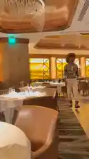 In a stylish restaurant with elegant chandeliers and warm lighting, a man named Delly confidently walks down the aisle. He is dressed in a patterned shirt, white pants, brown boots, and a wide-brimmed hat, showcasing his unique fashion sense. by @delwinbazilme with Ella Es Bonita, Donde Pisa Es Ley by Armand La Máquina