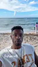 A man, identified as Delly, films himself on a sunny beach. He is wearing a white 'TEXAS' t-shirt and a chain necklace. The background shows the ocean, other people enjoying the water, and a clear blue sky. The text overlay reads '4:24 PM Intiernito'. The audio features the sound of crashing waves. by @delwinbazilme with Original Sound by delwinbazilme
