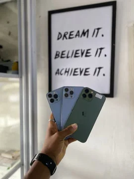 A hand holds three different colored iPhone 13 Pro Max models against a wall with a 'Dream It. Believe It. Achieve It.' poster. The scene is static, focusing on the phones' design and color options. by @ab_god18 with original sound