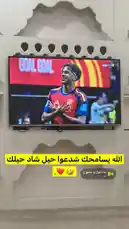 A television screen displays a football player celebrating a goal for the Spanish national team during a match. The word 'GOAL' is prominently shown on the screen. A voiceover in Arabic can be heard saying, 'يا زابح يا مزبوح' ('Oh butcher, oh slaughtered one'), followed by laughter. The setting appears to be indoors, with a decorative white wall behind the TV. by @snap_alshle with يا ذابح يا مذبوح by H
