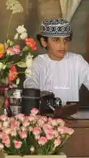 A young individual dressed in traditional Omani attire, including a white dishdasha and a black-and-white patterned kuma, poses thoughtfully beside vintage photography equipment and fresh floral arrangements. The scene captures cultural heritage, artistic expression, and timeless elegance, blending modern storytelling with classic style. by @nowaira-11 with original sound