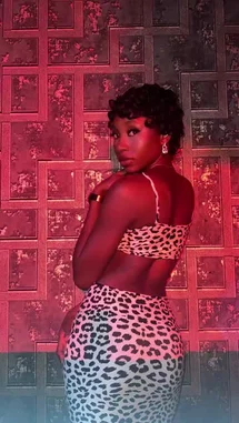 A woman with dark curly hair, wearing a striking leopard print two-piece outfit, poses confidently against a textured wall illuminated by dramatic red lighting. She turns her head over her shoulder to look directly at the camera, creating an intimate and powerful moment. A soulful female vocal performance plays in the background, adding emotional depth to the stylish visual. by @be-sassyish with Read All About It, Pt. III by Emeli Sandé