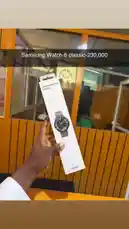 A hand holds up the retail packaging for the Samsung Galaxy Watch Classic, showcasing the product's sleek design and branding. The setting appears to be a modern office or tech store with glass partitions and wooden paneling in the background. by @frekasglobal with LET ME BE by The Second Voice