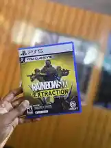 A close-up view of a person holding the physical copy of the PlayStation 5 game 'Tom Clancy's Rainbow Six Extraction'. The cover art features tactical operators in combat gear against a yellow and black background. The game case is clearly labeled for the PS5 console and published by Ubisoft. by @frekasglobal with original sound