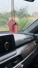 This slow-motion, 1 FPS video captures the serene interior of a car during a rainy day. Viewers see raindrops on the windshield, a stylish pink air freshener shaped like Africa hanging from the rearview mirror, and a modern dashboard display. The calm atmosphere is enhanced by Arabic text overlaying the image. This visually soothing clip invites curiosity about the car's design and the story behind the decor. by @om.louraa with original sound
