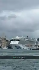 A stunning view of a massive cruise ship docked in a bustling harbor under a dramatic cloudy sky. The scene captures the grandeur of modern maritime travel juxtaposed with historic city architecture. A small boat navigates near the colossal vessel, emphasizing its scale. Arabic text overlay reads 'السعودية كروز اريبا' (Saudi Arabia Cruise Area), suggesting a regional travel context. The image evokes themes of adventure, luxury travel, and global exploration. by @n.c7x with original sound