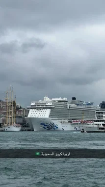 A stunning view of a massive cruise ship docked in a bustling harbor under a dramatic cloudy sky. The scene captures the grandeur of modern maritime travel juxtaposed with historic city architecture. A small boat navigates near the colossal vessel, emphasizing its scale. Arabic text overlay reads 'السعودية كروز اريبا' (Saudi Arabia Cruise Area), suggesting a regional travel context. The image evokes themes of adventure, luxury travel, and global exploration. by @n.c7x with original sound