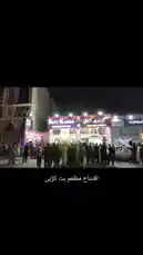 This nighttime scene captures the lively atmosphere outside 'Bitti Karan,' a bustling commercial hub. A large crowd gathers on the street, illuminated by the store's bright lights and festive decorations, creating a vibrant community gathering spot. by @om-5p with Original Sound by om-5p