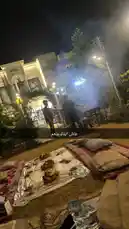 This captivating video captures a serene evening gathering hosted by Wasta Hawkar. Set against the backdrop of a beautifully lit house, the scene unfolds on a grassy lawn where guests relax on patterned rugs. A delicious spread of fresh fruits and snacks is artfully arranged on a tablecloth, inviting viewers to partake in the feast. In the background, a person tends to a sizzling barbecue grill, filling the air with tantalizing smoke and the promise of grilled delights. The warm glow of ambient lights creates an intimate and welcoming atmosphere, perfect for connecting with loved ones. This authentic slice-of-life moment highlights the simple pleasures of good food, company, and the gentle ambiance of a night spent outdoors. by @hawkar_service with original sound