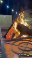 A close-up view of a vibrant outdoor fire burning brightly at night, creating a warm and inviting atmosphere. The flames dance over glowing embers and logs within a metal fire pit, set against a backdrop of string lights and a dark sky. This serene scene captures the essence of evening activities and community gatherings, likely part of the 'Kaizen Private Nursery & Evening Activities' program, showcasing a peaceful moment perfect for relaxation or socializing after dark. by @nouf4445 with جمعة احبابك by Contains: هذي الدنيا - غريب ال مخلص