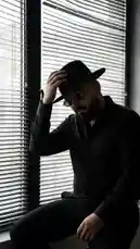 A man in all black, wearing a fedora and sunglasses, sits pensively by a window with blinds. The moody, cinematic shot captures a moment of quiet intensity, perfectly complemented by a soulful Arabic music track. by @djsefo with Ma3aya by Bahaa Sultan