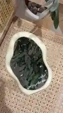 A close-up view of a uniquely shaped mirror with a cream-colored frame, resting on a woven rattan surface. The mirror reflects green foliage, creating a serene aesthetic. Arabic text is visible on the glass, adding a cultural design element. This stylish home decor piece is presented by متجر ريزن تبوك, showcasing modern interior design trends. by @r200an with original sound