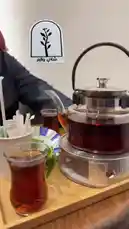 This close-up shot captures a beautifully arranged tea service, perfect for a relaxing moment. A traditional glass teapot filled with rich, dark Turkish tea sits atop a small electric warmer, keeping it hot. Beside it, a classic tulip-shaped glass is brimming with the same deep-colored brew. The entire setup rests on a warm wooden tray, creating an inviting and authentic atmosphere. This simple yet elegant scene evokes feelings of comfort and tranquility. by @azozi.5562 with original sound