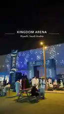 This nighttime scene captures the vibrant atmosphere at Kingdom Arena in Riyadh, Saudi Arabia. People dressed in traditional attire gather near illuminated entrance signs for 'Al Hilal' and 'Stairs,' suggesting a major event like a concert or sports game. The modern architecture and dynamic lighting create an energetic mood, offering a glimpse into the city's bustling entertainment district. by @abood114 with original sound