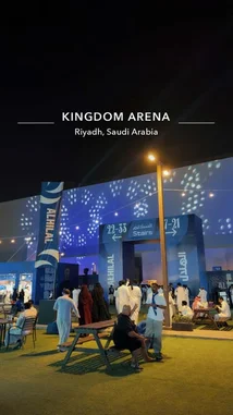 This nighttime scene captures the vibrant atmosphere at Kingdom Arena in Riyadh, Saudi Arabia. People dressed in traditional attire gather near illuminated entrance signs for 'Al Hilal' and 'Stairs,' suggesting a major event like a concert or sports game. The modern architecture and dynamic lighting create an energetic mood, offering a glimpse into the city's bustling entertainment district. by @abood114 with original sound