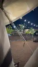 Experience the serene ambiance of a glamping site at night from the comfort of a tent. String lights illuminate the outdoor area, creating a warm and inviting atmosphere perfect for relaxation. This peaceful scene showcases a gravel ground, wooden benches, and young trees under a dark sky, ideal for those seeking a tranquil camping experience. by @karim.alsaleh with karim.alsaleh by Original Sound