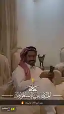 A man dressed in traditional Saudi attire, including a white thobe and red-and-white ghutra, smiles warmly while seated on a luxurious sofa. He holds a smartphone, seemingly sharing a moment of national pride. The background features elegant decor, including a prominent silver trophy, symbolizing achievement and heritage. This celebratory scene captures the essence of Saudi culture and patriotism. by @tr3i5 with لا استطيع by سنار