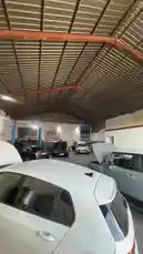 This time-lapse footage captures the bustling activity inside a professional auto repair garage. Multiple vehicles, including white and black sedans, are seen undergoing maintenance with hoods open under bright work lights. A large hydraulic lift stands ready in the background, while mechanics move about efficiently. The industrial space features a high, corrugated metal roof supported by red beams, highlighting the practical setup of a modern service center. by @hawkar_service with original sound