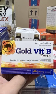 A close-up shot reveals a hand holding a box of Gold-Vit B Forte, a food supplement from Olymp Labs. The packaging highlights benefits for healthy skin, nervous system, metabolism, and blood. In the background, other health products like collagen and protein powder are visible, suggesting a focus on wellness and nutrition. This scene is ideal for triggering a deeper crawl due to its clear product focus and context. by @lanutritioniraq with original sound