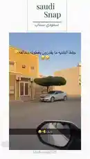 This short clip from 'Saudi Snap' showcases a silver car parked neatly in front of a distinctive yellow building with white window frames. A large, perfectly shaped tree stands prominently beside the vehicle, creating an unusual and photogenic composition. The scene is captured from a moving car, adding a dynamic feel. Arabic text overlays add a humorous or descriptive comment to the visual. by @saudi-snap2025 with Original Sound by saudi-snap2025