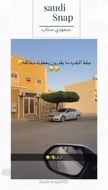 This short clip from 'Saudi Snap' showcases a silver car parked neatly in front of a distinctive yellow building with white window frames. A large, perfectly shaped tree stands prominently beside the vehicle, creating an unusual and photogenic composition. The scene is captured from a moving car, adding a dynamic feel. Arabic text overlays add a humorous or descriptive comment to the visual. by @saudi-snap2025 with Original Sound by saudi-snap2025