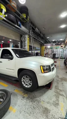 A white Chevrolet Suburban is parked inside a well-lit tire shop, positioned on a red hydraulic lift. The garage features extensive shelving stocked with tires and wheels, and a wall display of various rims. The scene is static, suggesting a focus on the vehicle and the shop's inventory. by @mo_kbs8 with 🖤🖤😻🤙🏻. by Sn: gk-n