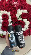A hand presents two black aerosol cans of Axe body spray, 'Axe Black' and 'Axe Alka,' against a luxurious backdrop of red and white roses. The DS Drogerie Store logo is visible, highlighting their selection of authentic German products. by @dsdrogeriestore with DS🖤❤️💛 by Ds drogerie store🇩🇪