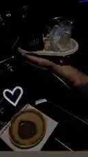 This intimate, dimly lit scene captures a relaxed moment inside a car. A hand holds a takeout cup of ice water next to a bottle of Cold Brew coffee. In the foreground, a small pastry with chocolate filling rests on a napkin, accompanied by a glowing white heart-shaped light. The overall atmosphere is calm and personal, suggesting a quiet break or a simple pleasure. by @tr3i5 with original sound