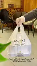 This video showcases a delivery moment from Makan Darb Al Hareer, highlighting their white branded takeout bag. A gloved hand holds the bag in a stylish restaurant setting, emphasizing hygiene and premium service. On-screen text includes contact details and a special offer: 'Raffle! Win a meal for free!' This scene promotes customer engagement and brand trust, ideal for attracting new patrons through social media. by @darbalhareer with original sound