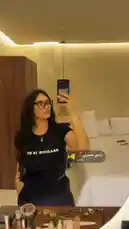 In this short clip, Soso Elewa takes a mirror selfie in a well-lit room. She is wearing a black t-shirt with the text 'READING IS HOT' printed on it, paired with dark pants. Her long, wavy hair and glasses are prominent features. The video has an Arabic music track playing in the background. by @codedsoso with رامي صبري❣️ by ازاييي