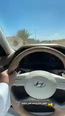 A driver's perspective view from inside a modern Hyundai vehicle, showcasing its luxurious interior, advanced digital dashboard, and steering wheel controls. The car is being driven on a sunny day through a suburban area with palm trees visible through the windshield. This immersive POV provides a real-world look at the vehicle's features and driving experience. by @n.c7x with اللهم صلّ وسلم على محمد by ..