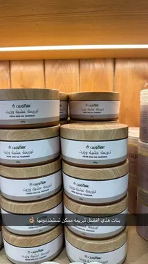This close-up video showcases stacks of 'Herb and Oil Taberna' body butter jars, highlighting their elegant wooden lids and minimalist design. A text overlay in Arabic promotes the product as a natural skincare solution. The warm lighting and wooden background create an inviting atmosphere, emphasizing the brand's focus on organic ingredients and traditional wellness. by @ru.ab71 with original sound
