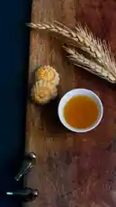 A serene top-down view captures a beautifully arranged still life perfect for autumn. On a warm, rustic wooden board, delicate mooncakes sit beside a small bowl of golden tea. Dried wheat stalks add a natural, seasonal touch. In the corner, elegant metallic tongs rest, hinting at a moment of quiet preparation. This peaceful scene evokes calmness and the simple pleasures of a traditional tea ceremony. by @moladone with original sound