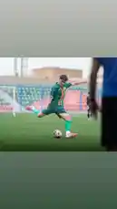 This dynamic clip captures a player from Al-Rawdah FC during an intense training session on a lush green pitch. Dressed in the team's signature green jersey with orange accents and matching socks, the athlete executes a powerful kick with focused precision. The stadium setting, complete with red seating and a visible goalpost, adds authenticity to the scene. With a teammate partially in frame and another player in the background, the atmosphere reflects real-world soccer drills and team camaraderie. by @alrawdahclub with original sound