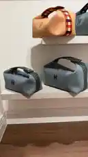 A stylish arrangement of designer handbags is showcased on minimalist white shelves against a clean wall. The collection features a variety of textures and colors, including a beige bag with a red-and-white checkered strap and two gray herringbone-patterned bags, each adorned with a prominent 'H' logo. The setting suggests a boutique or retail display, emphasizing modern elegance and fashion-forward design. by @lussowatches with original sound