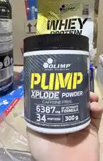 A close-up shot of a hand holding a container of Olimp Sport Nutrition's Pump Xplode Powder. The label clearly displays 'Caffeine Free' and '6387 mg of DetoNate Formula'. The product is presented as a pre-workout supplement with 34 portions and a net weight of 300g. In the background, other fitness supplements like whey protein are visible, suggesting a focus on sports nutrition. by @lanutritioniraq with original sound