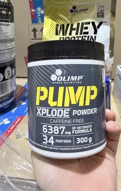 A close-up shot of a hand holding a container of Olimp Sport Nutrition's Pump Xplode Powder. The label clearly displays 'Caffeine Free' and '6387 mg of DetoNate Formula'. The product is presented as a pre-workout supplement with 34 portions and a net weight of 300g. In the background, other fitness supplements like whey protein are visible, suggesting a focus on sports nutrition. by @lanutritioniraq with original sound