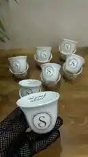 A hand wearing a black mesh glove carefully presents a set of pristine white ceramic coffee cups, each adorned with a monogrammed 'S' and Arabic calligraphy reading 'سيدة بن أسماء'. The cups are neatly stacked on a warm wooden table, suggesting a luxurious unboxing experience. This stylish display highlights personalized home goods perfect for gifting or self-care routines. by @r200an with original sound