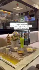 This video offers a close-up look at the interior of مطعم درب الحرير, showcasing their beautifully arranged table setting. Viewers can see a traditional golden Arabic coffee pot, delicate cups, a vibrant green beverage, and small bowls of snacks, all placed on an ornate table mat. In the background, glass display cases filled with desserts and drinks highlight the restaurant's offerings. The scene captures the ambiance and presentation style of this dining establishment. by @darbalhareer with Original Sound by darbalhareer
