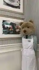 In this charming scene, Raghad presents a delightful setup featuring a life-sized teddy bear dressed as a café employee. The bear wears a crisp white apron with the 'Ralph Lauren' logo, standing beside a wall adorned with elegant framed pictures of a vintage green train and a coffee cup. The sophisticated interior, with its striped wallpaper and classic decor, creates a whimsical yet upscale atmosphere, blending playful design with high-end branding. by @i_vcil with original sound