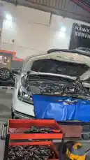 A static shot inside the Wasta Hawkar auto service garage. A white car with its hood open reveals the engine bay, protected by a blue cover. In the foreground, a red tool cart is filled with various tools. The background features a large banner with the 'HAWKAR SERVICE' logo, establishing the setting as a professional automotive repair shop. by @hawkar_service with Original Sound by hawkar_service