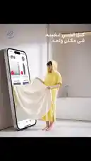 In this innovative scene, RJ demonstrates a cutting-edge marketing concept. A woman in a yellow hooded robe stands beside a massive smartphone screen displaying a product page. She holds up a cream-colored towel, seemingly pulling it out of the digital world into reality. The Arabic text 'كل اللي تبغينه في مكان واحد' translates to 'Everything you want in one place,' highlighting the seamless integration of online shopping and physical experience. This visually striking moment is designed to capture attention and encourage viewers to explore RJ's unique offerings. by @rjrobes with original sound