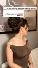 A woman with dark hair styled in an elegant, voluminous updo is shown from the side. She wears a brown, off-the-shoulder dress with gold ring details and a sparkling choker necklace. An Arabic text overlay expresses admiration for her beauty and style. The background features a large framed picture of a mountain landscape. by @rii.38 with يا اغلى من روحي علي 🤍 by My favvv