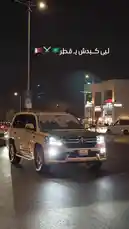 A luxury beige Toyota Land Cruiser drives through a city street at night, its headlights illuminating the wet pavement. The vehicle features a large graphic on the windshield depicting men in traditional Saudi thobes and ghutras, suggesting a cultural or celebratory theme. The urban background includes illuminated storefronts and streetlights, creating a vibrant nighttime atmosphere. by @wedad_23g with في قطر نحيا ونفنى by 🇶🇦