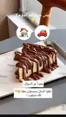 A close-up shot of a decadent slice of cheesecake drizzled with chocolate sauce sits on a metal tray. Text overlays in Arabic pose a playful dilemma: 'Which do you choose?' with icons of a house and a car. The scene includes a 'souvenir' branded paper liner, suggesting a café setting. This visually rich, question-driven moment is designed to spark engagement and encourage viewers to make a choice—perfect for driving interaction and triggering deeper exploration. by @sequence-coffee with original sound