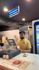 In this behind-the-scenes look at the 'الرامــال-restaurant' counter, a staff member demonstrates a calm and professional approach to a customer interaction. The scene captures the everyday operations of a modern restaurant, highlighting effective communication and service. by @sandshawarma with Original Sound by sandshawarma