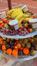 A vibrant, two-tiered platter filled with fresh fruits like bananas, strawberries, grapes, oranges, kiwis, and plums, artfully arranged around jars of Nutella. This appetizing setup suggests a delightful snack or dessert option, possibly showcased during evening activities at Kaizen Rawaqa Alia. The colorful presentation highlights healthy ingredients combined with indulgent chocolate spread, perfect for gatherings or family events. by @nouf4445 with original sound