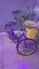 A vibrant purple bicycle adorned with a wicker basket full of colorful flowers is displayed indoors under soft purple lighting. The bike is positioned next to a large green plant against a textured wall, creating a stylish and decorative scene. This static shot captures a charming blend of urban cycling aesthetics and home decor. by @aded.ksa with original sound